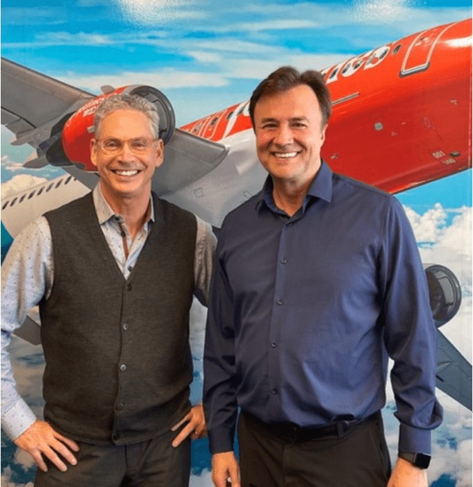 two men smiling big infront of a photo of a plane