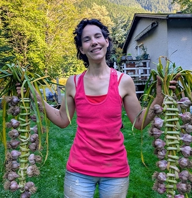 A woman smiling holding large strands of garlic from her garden