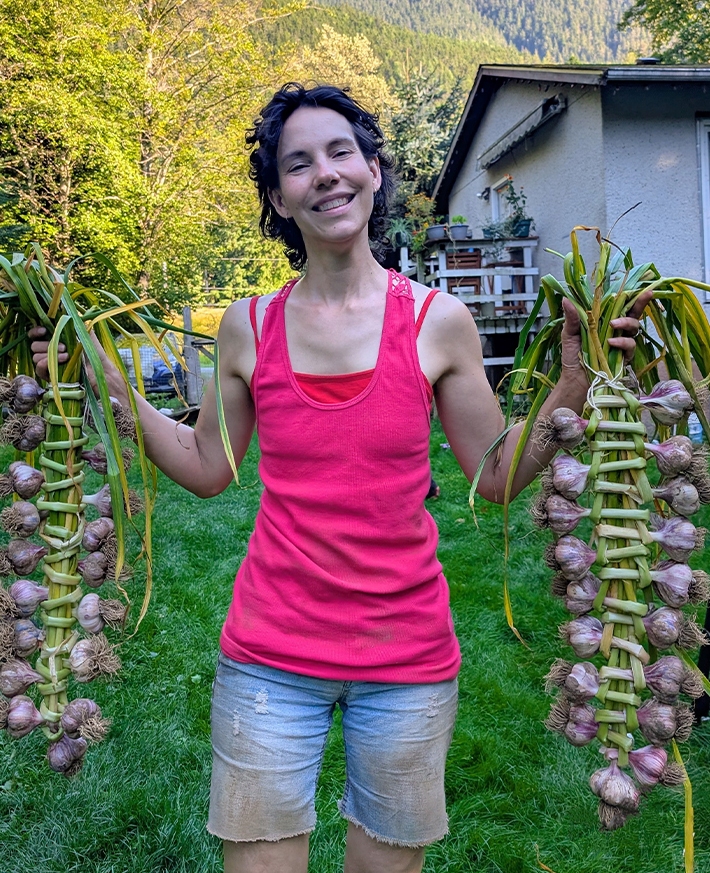 A woman smiling holding large strands of garlic from her garden